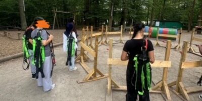 Mehrere Mädchen stehen vor einzelnen kleinen Holzzäunen. Sie tragen Kletterausrüstung und scheinen die ersten Schritte des Absicherns beim Klettern zu lernen. Das ganze findet draußen in einem Kletterwald statt.