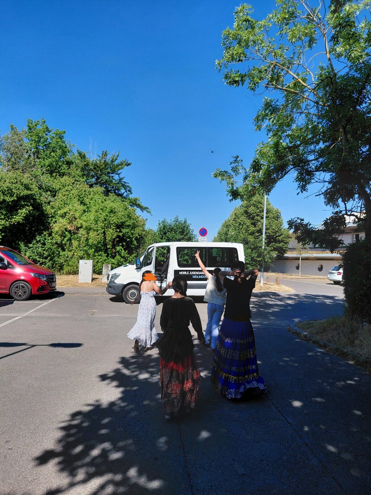Eine Gruppe von vier Frauen, die traditionelle Kleidung tragen, geht auf einer Straße. Hinter ihnen steht ein weißer Kleinbus. Auf der linken Seite ist ein roter Van geparkt.