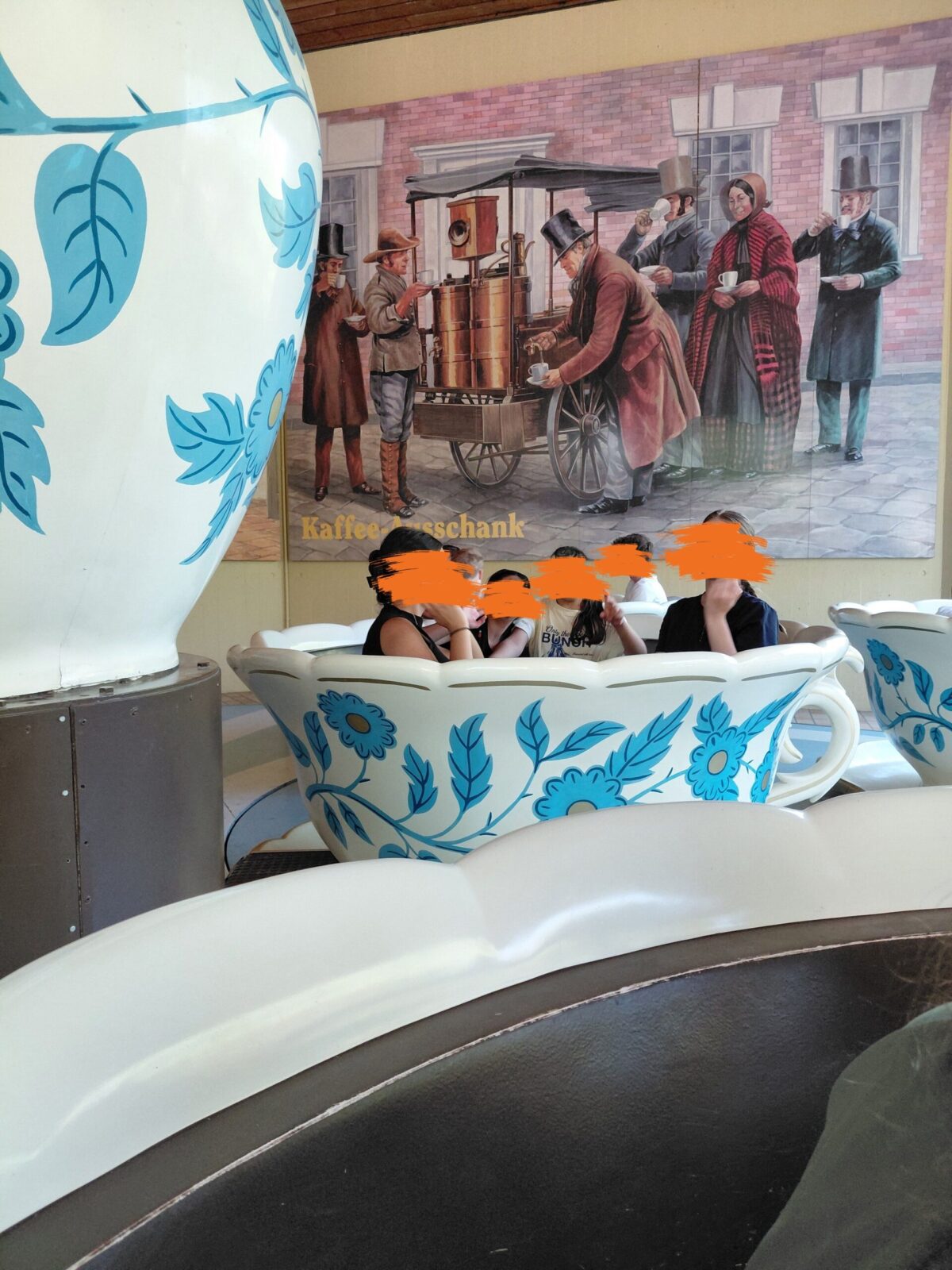 Eine Gruppe von Menschen sitzt in einer großen Tasse mit blauen Blumen. Die Tasse steht auf einem Tisch. Hinter ihnen ist ein großes Plakat mit Menschen in historischen Kostümen, die Kaffee trinken.