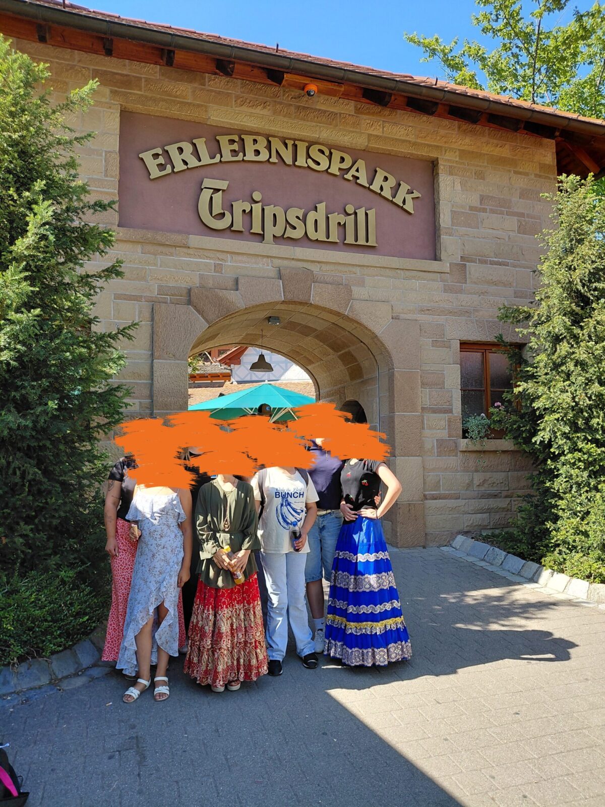 Eine Gruppe von Menschen steht vor dem Eingang des "Erlebnisparks Tripsdrill". Einige tragen traditionelle Kleidung. Der Eingang ist mit einem Bogen und einem Schild gestaltet.