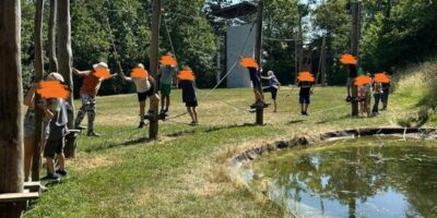 Eine Gruppe von Kindern, deren Gesichter unkenntlich gemacht wurden, nimmt auf einem Spielplatz an einer Seilbahn-Aktivität teil. Die Kinder stehen auf Plattformen, die durch Seile verbunden sind, und scheinen sich auf die Übung vorzubereiten. Im Hintergrund ist ein Teich zu sehen, der von Gras und Bäumen umgeben ist, was auf eine natürliche und entspannende Umgebung hinweist.