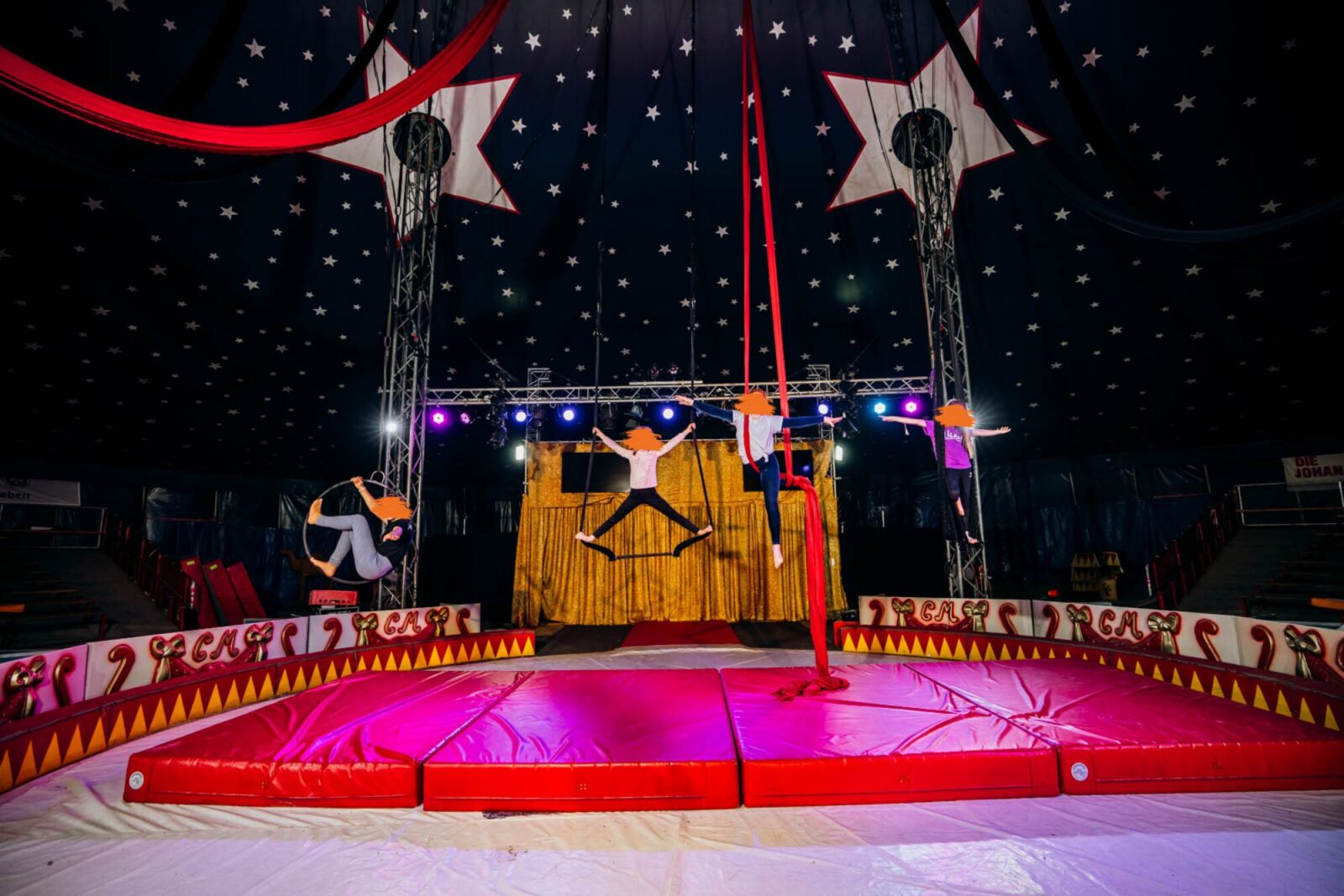 Vier junge Mädchen trainieren gemeinsam in der Zirkusmanege an verschiedenen Tüchern und Ringen. Die Kulisse ist eine goldene Bühne mit einem sternenübersäten Zirkuszeltdach. Die Atmosphäre wirkt lebendig und dynamisch, mit farbigem Bühnenlicht.