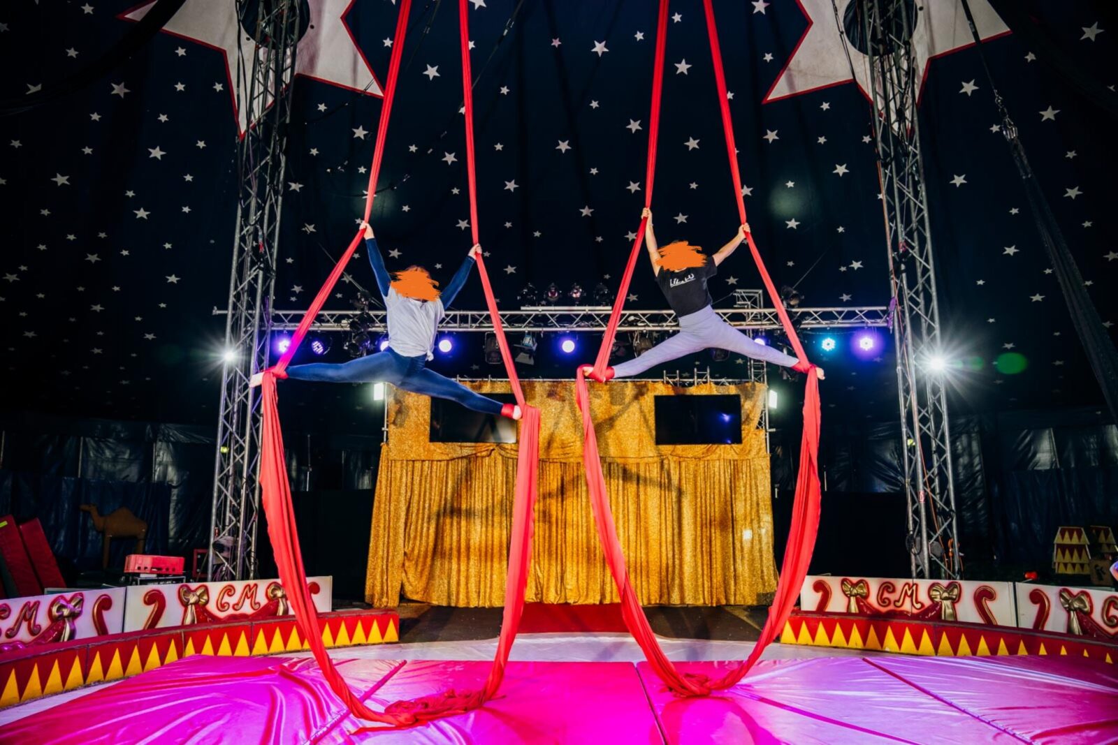 Zwei Mädchen zeigen eine synchronisierte Spagat-Pose an roten Tüchern in der Manege. Die Bühne im Hintergrund ist mit goldfarbenen Vorhängen geschmückt, und die Decke ist mit Sternen dekoriert. Die Szene vermittelt Eleganz und kraftvolle Körperbeherrschung.