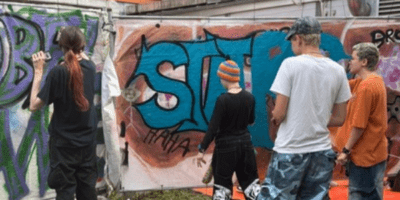 Eine Gruppe von Menschen unterschiedlichen Alters steht vor einer bunten Graffiti-Wand und betrachtet die kunstvollen Street-Art-Werke mit großflächigen Schriftzügen in Blau, Grün und Lila. Die Szene spielt sich in einem urbanen, von Industriegebäuden umgebenen Umfeld ab und zeigt eine gemeinschaftliche Aktivität im Bereich Urban Art. Die Teilnehmer wirken engagiert und interessiert an der kreativen Kunstform, die das Bild ausstrahlt.