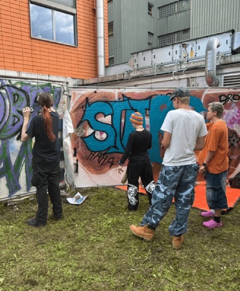 Eine Gruppe von Menschen unterschiedlichen Alters steht vor einer bunten Graffiti-Wand und betrachtet die kunstvollen Street-Art-Werke mit großflächigen Schriftzügen in Blau, Grün und Lila. Die Szene spielt sich in einem urbanen, von Industriegebäuden umgebenen Umfeld ab und zeigt eine gemeinschaftliche Aktivität im Bereich Urban Art. Die Teilnehmer wirken engagiert und interessiert an der kreativen Kunstform, die das Bild ausstrahlt.