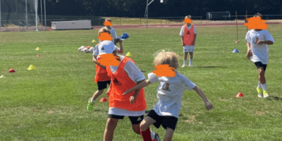 Mehrere Kinder spielen bei sonnigem Wetter Fußball auf einem grünen Rasenplatz während eines Trainings. Im Vordergrund kämpfen zwei Jungen intensiv um den Ball, wobei einer ein orangefarbenes Leibchen und der andere ein weißes T-Shirt trägt. Im Hintergrund sind weitere Mitspieler in Sportkleidung, Trainingshütchen, Tore und eine dichte Baumreihe zu sehen.