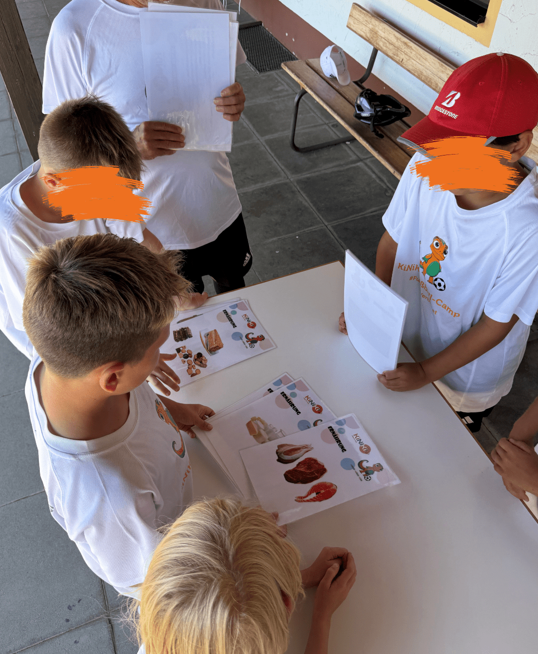 Eine Gruppe von Kindern steht im Freien um einen Tisch herum und schaut sich Informationsblätter zum Thema Ernährung an. Die Blätter zeigen das Logo „KINI KI“ und verschiedene Abbildungen von Lebensmitteln wie Fleisch, Fisch und Süßigkeiten. Ein Erwachsener im Hintergrund hält ebenfalls Dokumente in den Händen, während die Kinder in weißen T-Shirts aufmerksam den Inhalt studieren.