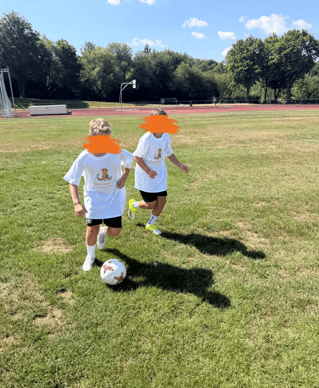 Zwei Jungen in weißen T-Shirts mit einem kleinen Dinosaurier-Logo laufen bei Sonnenschein über einen grünen Fußballplatz. Einer der Jungen führt den Fußball eng am Fuß, während der andere dicht neben ihm herläuft. Im Hintergrund erstreckt sich eine rote Laufbahn, ein Basketballkorb sowie eine dichte Reihe grüner Bäume unter blauem Himmel.