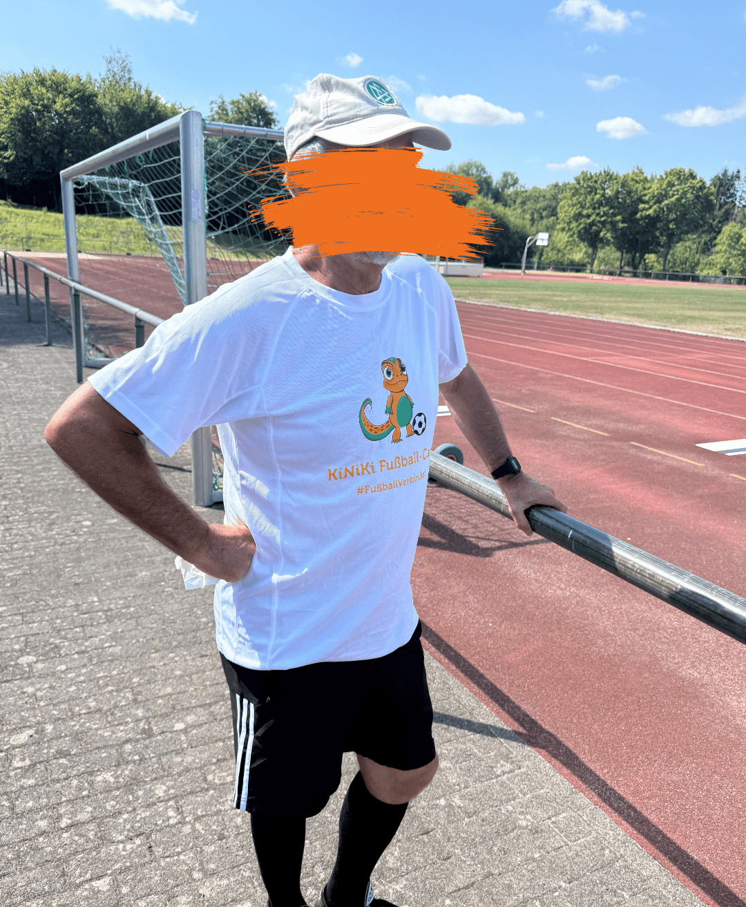 Ein älterer Mann mit einer hellen Kappe steht an einem Geländer neben einer roten Laufbahn auf einem Sportplatz. Er trägt ein weißes T-Shirt mit der Aufschrift „KiNiKi Fußball-Camp“ und einem kleinen Dinosaurier-Motiv sowie eine schwarze Sporthose. Im Hintergrund sind ein Fußballtor mit Netz, eine Laufbahn und grüne Bäume unter leicht bewölktem Himmel zu sehen.