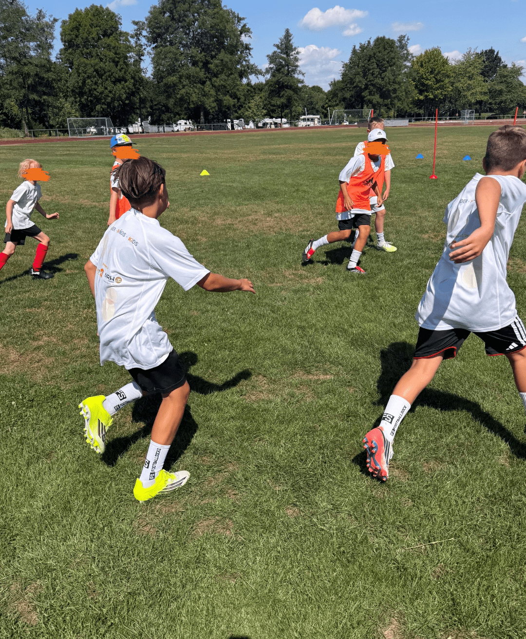 Mehrere Kinder laufen während eines Fußballtrainings dynamisch über einen grünen Rasenplatz. Einige Spieler tragen weiße T-Shirts mit Sponsorenaufdrucken, während andere in orangefarbenen Leibchen zu sehen sind. Im Hintergrund erstrecken sich kleine Trainingshütchen, rote Slalomstangen sowie eine weitläufige Reihe grüner Bäume unter strahlend blauem Himmel.