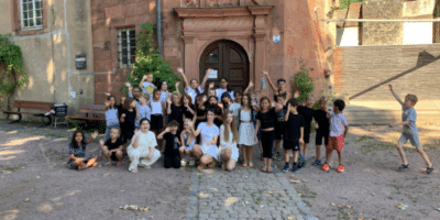 Eine große Gruppe von Kindern und Betreuern posiert fröhlich auf dem gepflasterten Innenhof vor dem Eingang eines historischen Gebäudes. Einige Kinder knien vorne, während die anderen dahinter stehen und jubelnd die Hände in die Luft strecken. Das Bild zeigt die Gemeinschaft und gute Stimmung aller Teilnehmenden des Musikcamps im Freien.