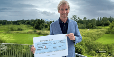 Ein Mann in einem blauen Sakko steht auf einer Terrasse mit Blick auf eine grüne Landschaft und hält einen großen symbolischen Scheck in den Händen. Der Scheck stammt von der Charitybank GmbH und zeigt einen Betrag von 2.160 Euro, der für einen wohltätigen Zweck bestimmt ist. Im Hintergrund sind Bäume, Wiesen und ein bewölkter Himmel zu sehen.