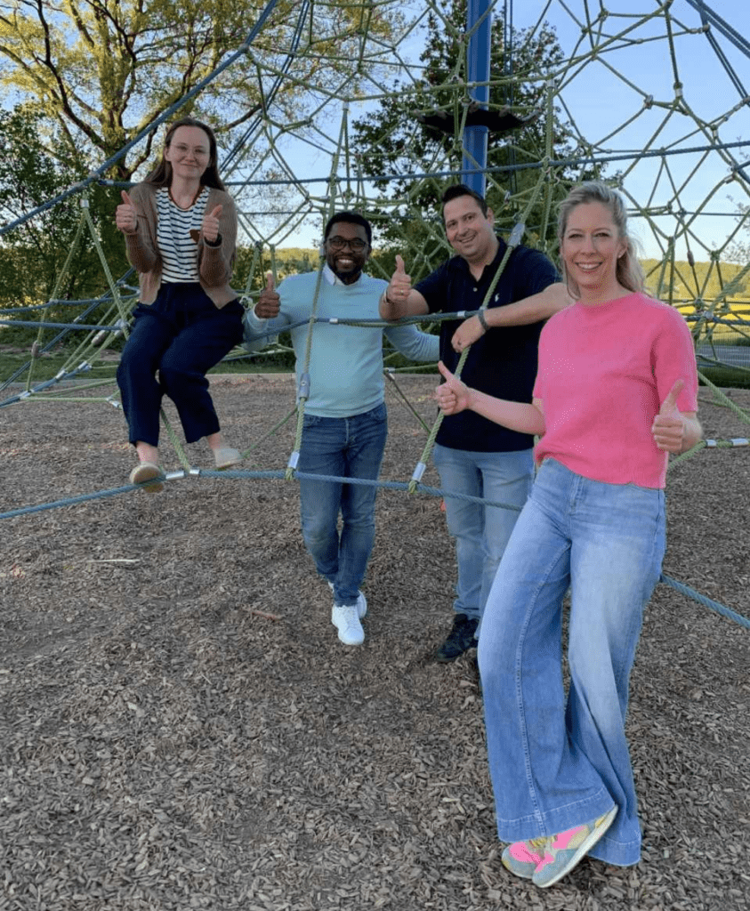 Gruppenfoto von fünf Personen unterschiedlichen Alters, die fröhlich zusammen in einer geodätischen Kuppelstruktur auf einem Spielplatz stehen und Daumen hoch zeigen. Die Gruppe lächelt in die Kamera und wirkt freudvoll und verbunden. Im Hintergrund sind grüne Bäume und ein blauer Himmel zu sehen, was auf ein Außengelände oder einen Park hindeutet.