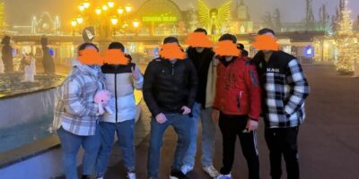 Eine Gruppe von sechs Personen steht nebeneinander vor einem Gebäude mit heller Beleuchtung. Ihre Gesichter sind unkenntlich gemacht.