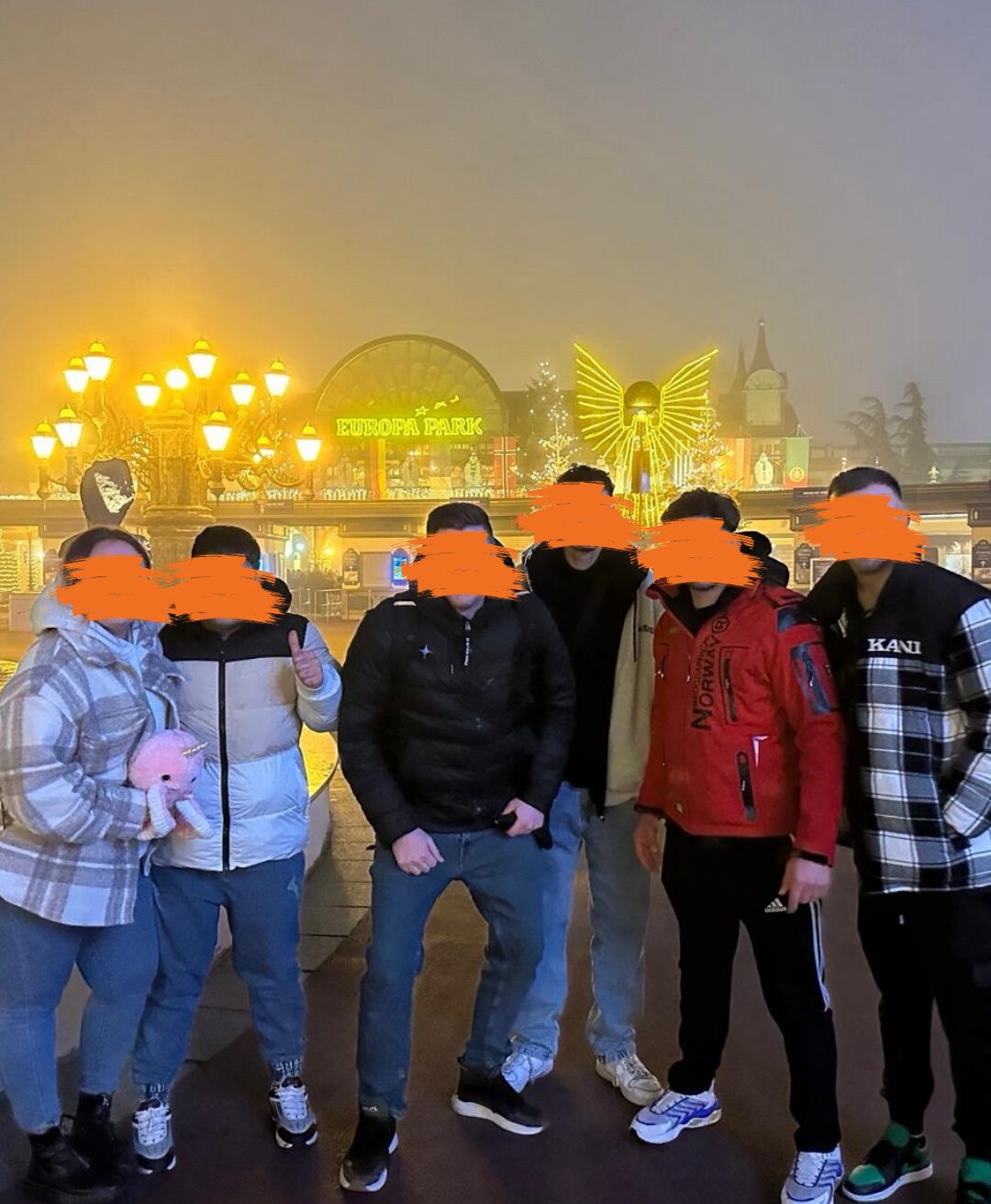 Eine Gruppe von sechs Personen steht nebeneinander vor einem Gebäude mit heller Beleuchtung. Ihre Gesichter sind unkenntlich gemacht.