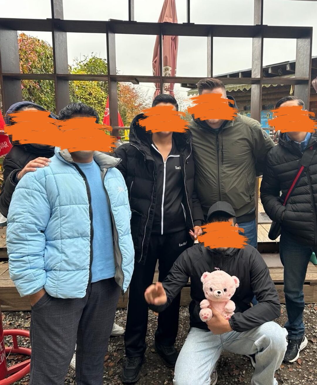 Eine Gruppe von sechs Personen steht nebeneinander, einer hockt und hält ein rosa Teddybär. Ihre Gesichter sind unkenntlich gemacht.