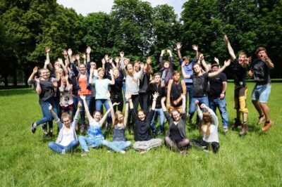 Eine große Gruppe von Menschen steht auf einer grünen Wiese und springt in die Luft. Sie wirken fröhlich und energetisch, während sie ihre Arme in die Höhe strecken. Im Hintergrund sind üppige, grüne Bäume zu sehen, die eine entspannte und natürliche Atmosphäre schaffen.