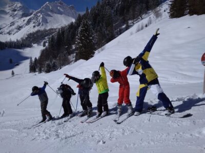 Eine Gruppe von sechs Skifahrern steht nebeneinander auf einer verschneiten Piste, umgeben von schneebedeckten Bergen und Bäumen. Sie tragen bunte Winterkleidung und Skiausrüstung, während sie fröhlich posieren und einige den Dab-Tanz machen. Die Sonne scheint hell und der Himmel ist klar, was zu einer perfekten Skiumgebung beiträgt.