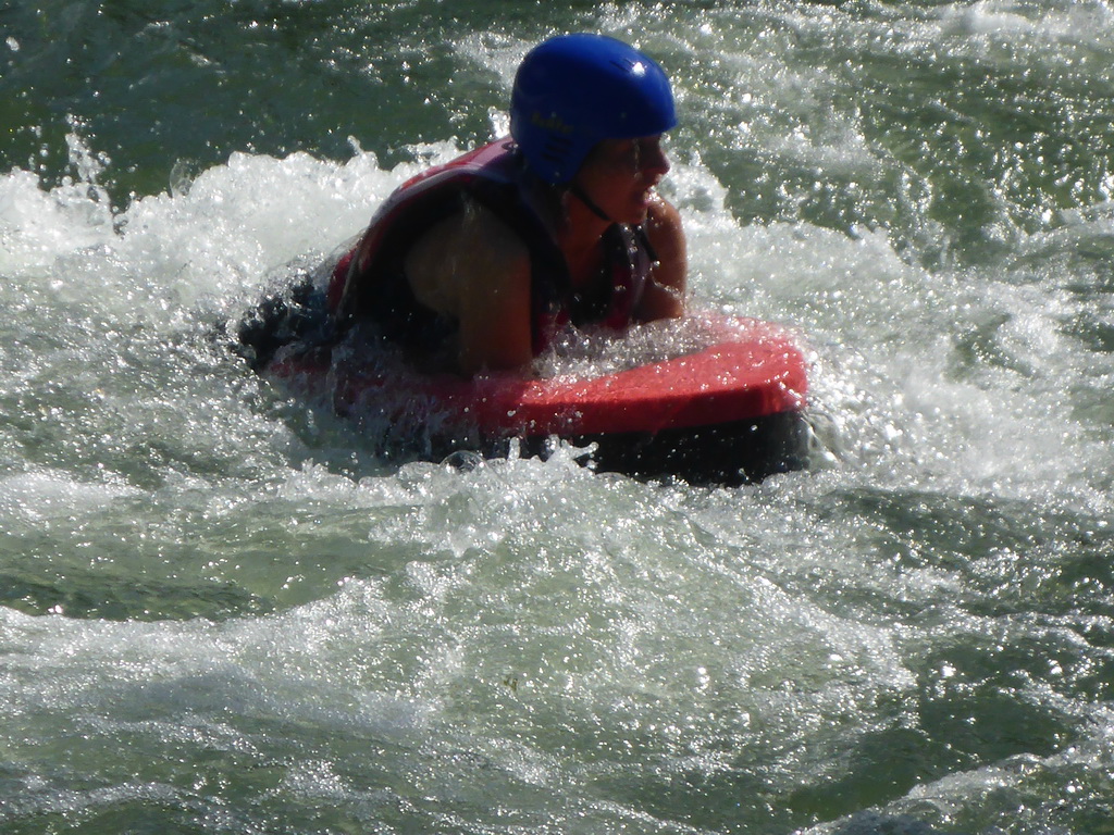 Eine Person trägt einen blauen Helm und liegt auf einem roten Schwimmbrett im Wasser. Die Szene zeigt spritzendes Wasser und deutet auf eine actionreiche Aktivität hin. Die Person scheint sich in einem Wildwasser zu befinden und genießt das Abenteuer.
