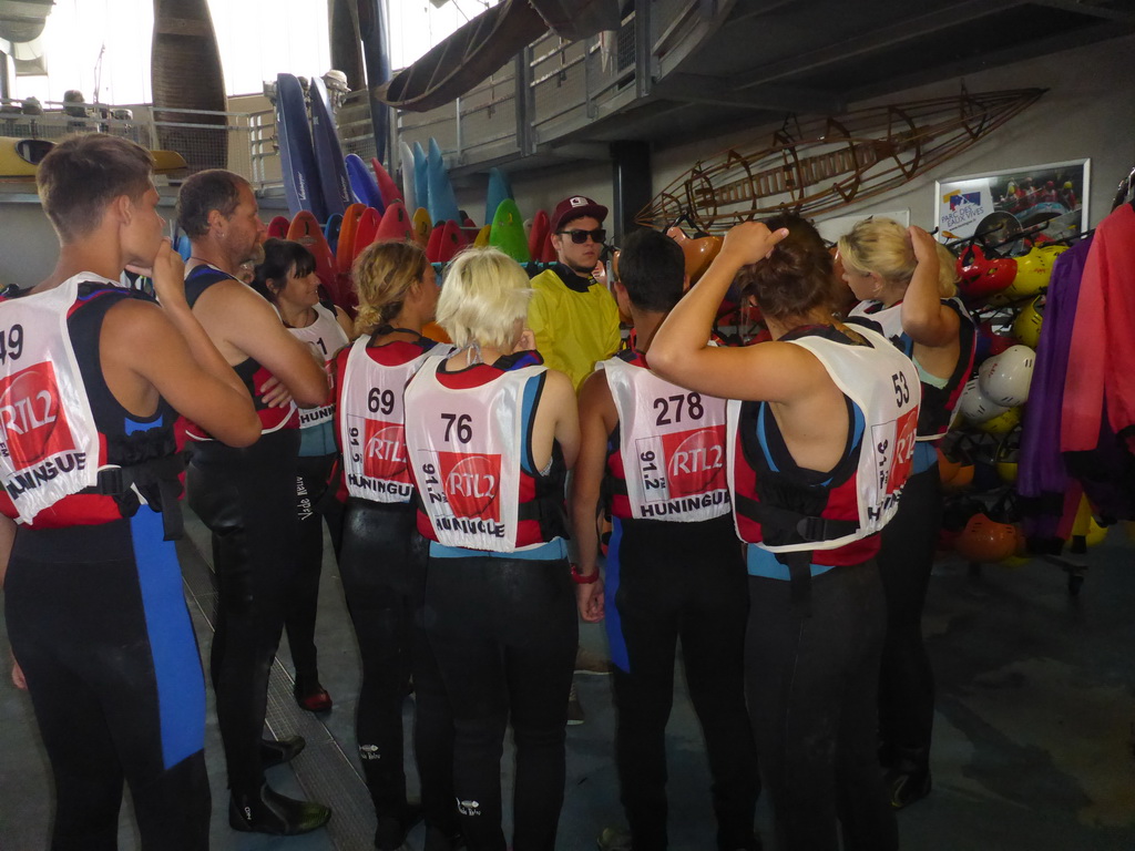 Eine Gruppe von Menschen steht in einem Indoor-Kajakzentrum und trägt Schwimmwesten mit aufgedruckten Nummern. Sie bereiten sich auf eine Wasseraktivität vor und scheinen Anweisungen zu erhalten. Im Hintergrund sind bunte Kajaks und verschiedene Ausrüstungsgegenstände zu sehen.