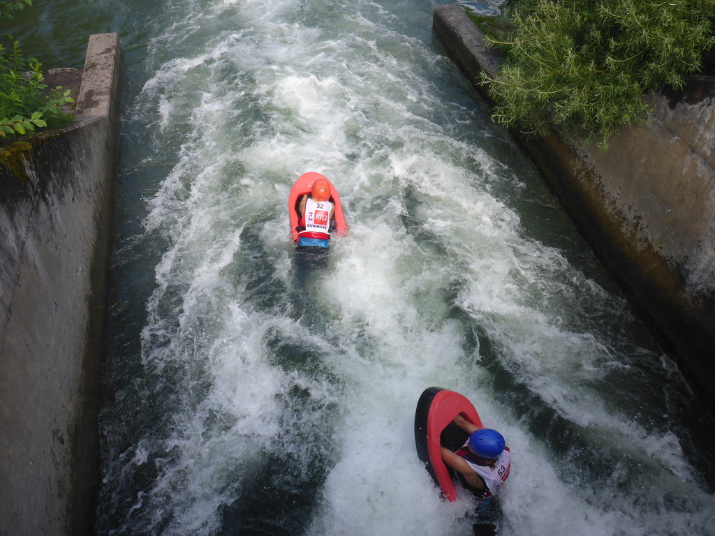 Zwei Personen treiben in einem engen Kanal mit stark strömendem Wasser auf Rettungsringen. Beide tragen Helme und Schwimmwesten, während sie die turbulente Fahrt genießen. Die Umgebung ist von Betonwänden und grüner Vegetation gesäumt, die das Abenteuer noch aufregender machen.