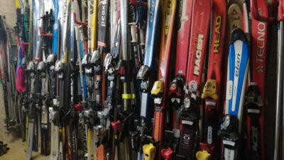 In diesem Bild sind viele Skier unterschiedlicher Marken und Modelle zu sehen, die ordentlich nebeneinander aufgereiht sind. Die Skier sind vertikal an einer Wand befestigt und zeigen eine Vielzahl von Farben und Designs. Im Hintergrund sind auch einige Skistöcke zu erkennen, die ebenfalls in einer geordneten Weise gelagert sind.