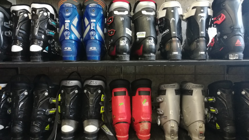 Eine Reihe von Skischuhen, die ordentlich auf Regalen gestapelt sind. Die obere Reihe zeigt verschiedene Modelle und Farben, darunter blaue, schwarze und rote Skischuhe. Die untere Reihe besteht ebenfalls aus verschiedenen Skischuhen, darunter schwarze, rote und graue Modelle, die für den Einsatz im Wintersport bereitstehen.