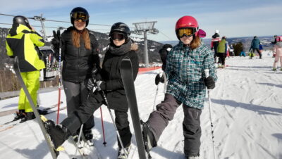 Eine Gruppe von Skifahrern steht auf einer verschneiten Piste und posiert fröhlich für ein Foto. Die Skifahrer tragen warme Winterbekleidung und Helme, bereit für eine Abfahrt. Im Hintergrund sind ein Skilift und andere Wintersportler zu sehen, die das sonnige Wetter genießen.