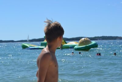 Ein Junge steht am Strand und schaut aufs Meer hinaus. Im Wasser sind mehrere Menschen, die sich im Meer vergnügen. Im Hintergrund sind große aufblasbare Wasserspielzeuge zu sehen, die auf dem Wasser treiben. Die Sonne scheint hell und der Himmel ist klar und blau.
