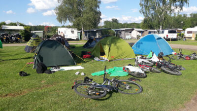 Ein Campingplatz mit mehreren Zelten auf einer grünen Wiese. Fahrräder liegen verstreut vor den Zelten, und Campingausrüstung ist auf dem Boden verteilt. Im Hintergrund sind Wohnwagen und einige Bäume zu sehen. Der Himmel ist klar und sonnig mit wenigen Wolken.