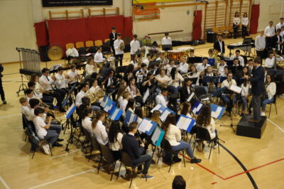 Ein großes Orchester, bestehend aus jungen Musikerinnen und Musikern, spielt in einer Turnhalle. Der Dirigent steht auf einem Podest und leitet das Ensemble, während die Musiker konzentriert ihre Instrumente spielen. Im Hintergrund sind verschiedene Schlaginstrumente und weitere Musiker zu sehen, die ebenfalls Teil des Orchesters sind.
