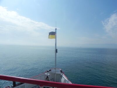 Das Bild zeigt den Bug eines Bootes auf offenem Wasser, bei klarem Himmel und ruhiger See. Eine Flagge, die in den Farben Schwarz und Gelb gehalten ist, weht an einem Mast am Vorderdeck des Bootes. Im Hintergrund erstreckt sich der Horizont, an dem das Wasser und der Himmel aufeinandertreffen, während einige Segelboote in der Ferne zu sehen sind.