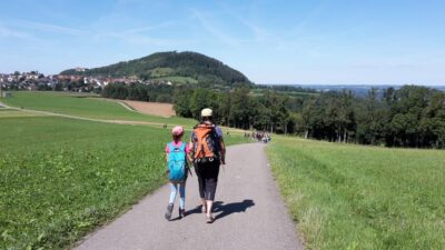 Eine Gruppe von Wanderern geht auf einem asphaltierten Weg durch eine grüne Landschaft. Im Vordergrund sind zwei Personen zu sehen, die Wanderrucksäcke tragen und miteinander sprechen. Im Hintergrund erstreckt sich ein Dorf mit Häusern und ein bewaldeter Hügel unter einem klaren, blauen Himmel.