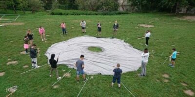 Eine Gruppe von Kindern und Jugendlichen steht im Kreis um eine große Plane auf einer Wiese. Sie bereiten sich darauf vor, die Plane aufzubauen, indem sie Stangen und andere Materialien halten. Die Szene deutet auf eine Outdoor-Aktivität oder ein gemeinschaftliches Projekt hin, möglicherweise ein Zeltaufbau oder ein Gruppenspiel im Freien.