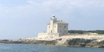 Das Bild zeigt einen historischen Leuchtturm auf einer felsigen Küste, umgeben von klarem, blauem Wasser. Der Leuchtturm ist aus hellem Stein gebaut und hat ein klassisches Design mit einem weißen Lichtturm an der Spitze. Im Hintergrund ist eine bewaldete Landschaft zu sehen, die den friedlichen und abgeschiedenen Charakter des Ortes unterstreicht.
