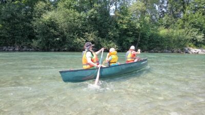 Drei Personen paddeln in einem grünen Kanu auf einem klaren Fluss. Alle tragen orangefarbene Schwimmwesten und genießen die sonnige Umgebung. Im Hintergrund sind üppige grüne Bäume und Sträucher zu sehen, die das Ufer des Flusses säumen.