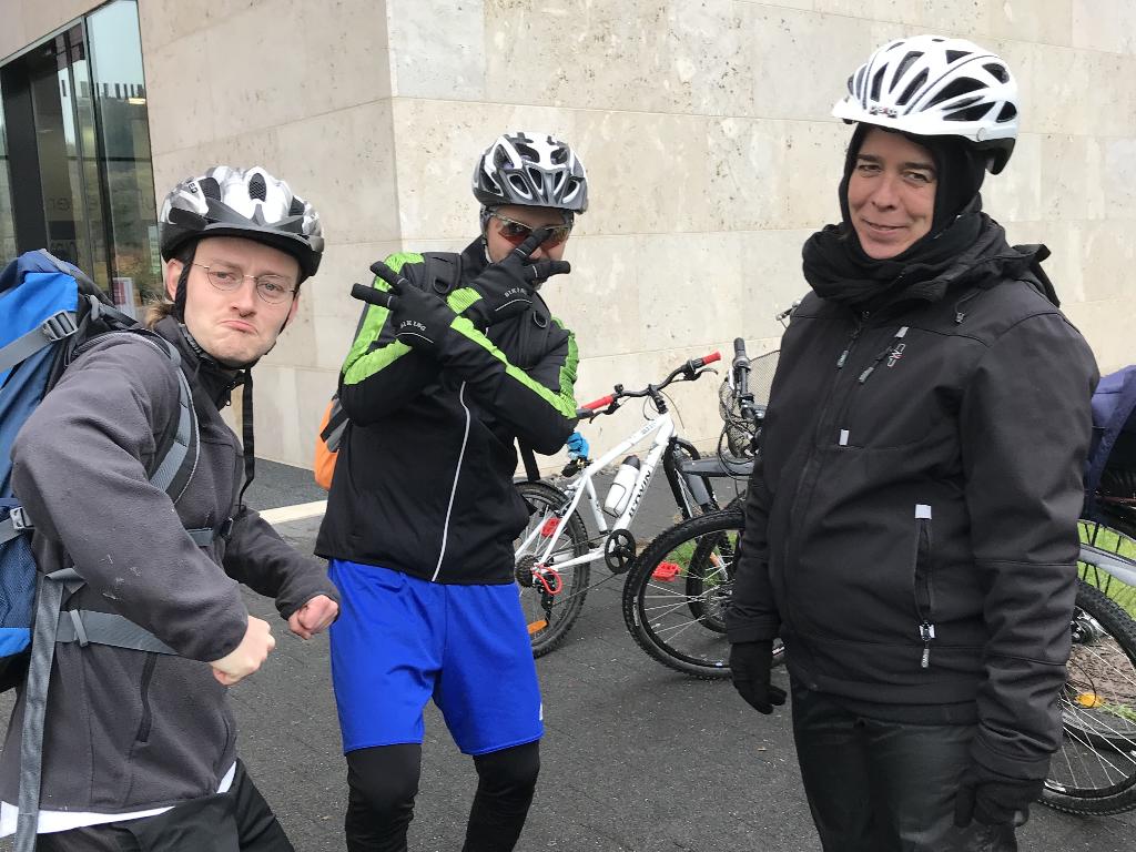 Drei Personen stehen in Fahrradkleidung und Helmen vor einem Gebäude. Im Hintergrund sind mehrere Fahrräder zu sehen, die an einem Fahrradständer abgestellt sind. Die Personen tragen wetterfeste Kleidung und scheinen bereit für eine Fahrradtour zu sein.