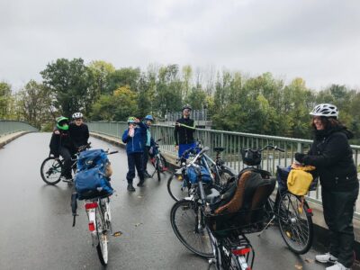 Eine Gruppe von Radfahrern macht eine Pause auf einer Brücke. Die Fahrräder sind mit Gepäcktaschen und Körben ausgestattet, die auf eine längere Tour hinweisen. Im Hintergrund sind Bäume zu sehen, die auf einen herbstlichen Tag schließen lassen.