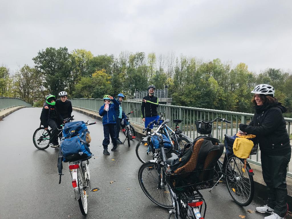 Eine Gruppe von Radfahrern macht eine Pause auf einer Brücke. Die Fahrräder sind mit Gepäcktaschen und Körben ausgestattet, die auf eine längere Tour hinweisen. Im Hintergrund sind Bäume zu sehen, die auf einen herbstlichen Tag schließen lassen.
