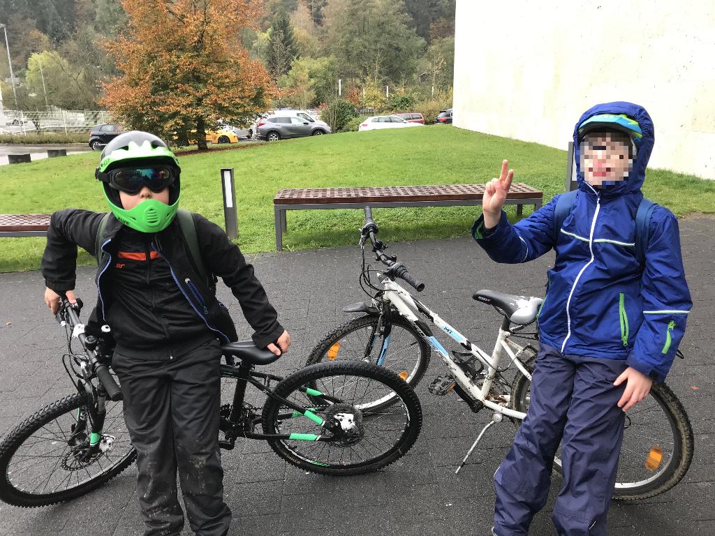 Zwei Kinder stehen neben ihren Fahrrädern auf einem gepflasterten Platz. Beide tragen wetterfeste Kleidung und Helme, bereit für eine Fahrradtour. Im Hintergrund sind grüne Rasenflächen, einige Bäume mit Herbstlaub und parkende Autos zu sehen.