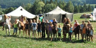 Eine Gruppe von Menschen steht auf einer Wiese vor mehreren großen Zelten. Einige Personen halten Pferde, während andere neben ihnen stehen. Im Hintergrund sind Bäume und Hügel zu sehen, die eine malerische Landschaft bilden.
