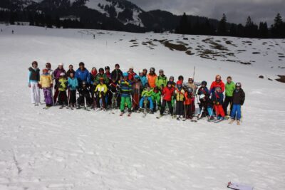 Eine große Gruppe von Skifahrern steht auf einer verschneiten Piste und posiert für ein Foto. Die Personen tragen bunte Skibekleidung und Skiausrüstung. Im Hintergrund sind schneebedeckte Berge und Bäume zu sehen, die eine winterliche Landschaft darstellen.