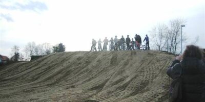 Eine Gruppe von Fahrradfahrern steht auf einem Hügel und bereitet sich auf eine Fahrt vor. Im Vordergrund sieht man eine Person, die die Szene fotografiert. Der Himmel ist bewölkt und die Umgebung ist von kahlen Bäumen umgeben, was auf eine kühle Jahreszeit hinweist.