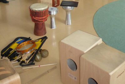 Das Bild zeigt eine Sammlung von Musikinstrumenten, die auf einem Boden arrangiert sind. Zu sehen sind unter anderem eine Trommel, eine Darbuka, ein Tamburin und mehrere Cajóns. Zudem ist eine Tasche mit verschiedenen Drumsticks und Percussion-Instrumenten zu erkennen.