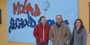Drei Personen stehen vor einer bunten Wand mit Graffiti, die die Worte "Mode" und "Agenda" zeigt. Die Wand ist in kräftigen Farben gestaltet und zieht die Aufmerksamkeit auf sich. Die Personen tragen warme Winterkleidung, was auf kaltes Wetter hinweist.