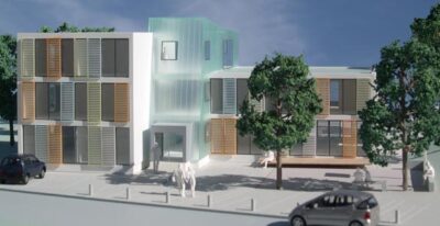Das Bild zeigt ein modernes Architekturmodell eines Gebäudes mit einer Kombination aus Glas- und Holzfassaden. Das Zentrum des Gebäudes ist mit transparentem Glas gestaltet, das Licht durchlässt und einen futuristischen Eindruck vermittelt. Auf der linken und rechten Seite befinden sich zwei Flügel mit farbigen Lamellen und großen Fenstern. Vor dem Gebäude sind einige Bäume und Autos zu sehen, die eine urbane Umgebung andeuten.