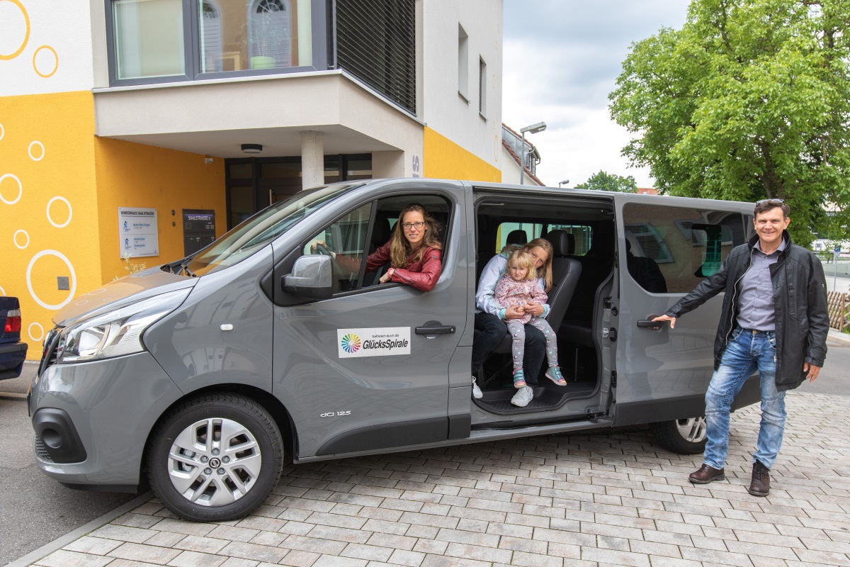 Ein grauer Kleinbus steht vor einem modernen Gebäude mit gelber und weißer Fassade. Der Kleinbus trägt das Logo der GlücksSpirale auf der Seite. Mehrere Personen sitzen im Fahrzeug, während eine Person draußen neben der geöffneten Tür steht.