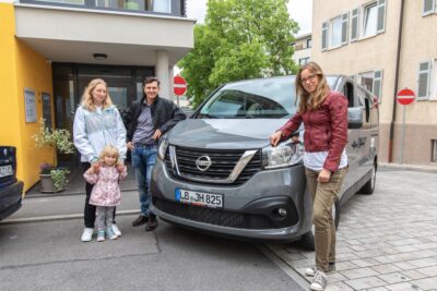 Eine Familie steht neben einem grauen Nissan-Van auf einer Straße. Im Hintergrund ist ein gelbes Gebäude mit Pflanzen und Schildern zu sehen. Die Szene wirkt alltäglich und zeigt die Familie in einer urbanen Umgebung.