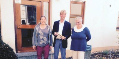 Drei Personen stehen vor einem Gebäude mit einer Holztür und Fenstern. Eine Person in der Mitte hält ein Buch oder Dokument in den Händen. Die Szene wirkt wie ein Treffen oder eine kleine Versammlung vor einem öffentlichen oder privaten Gebäude.