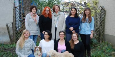Eine Gruppe von neun Frauen posiert für ein Foto in einem Garten. Einige von ihnen sitzen auf dem Boden, während andere stehen hinter ihnen. Zwei Hunde sind ebenfalls Teil des Bildes und sitzen neben den Frauen. Im Hintergrund sind ein Holzgitter und grüne Pflanzen zu sehen.